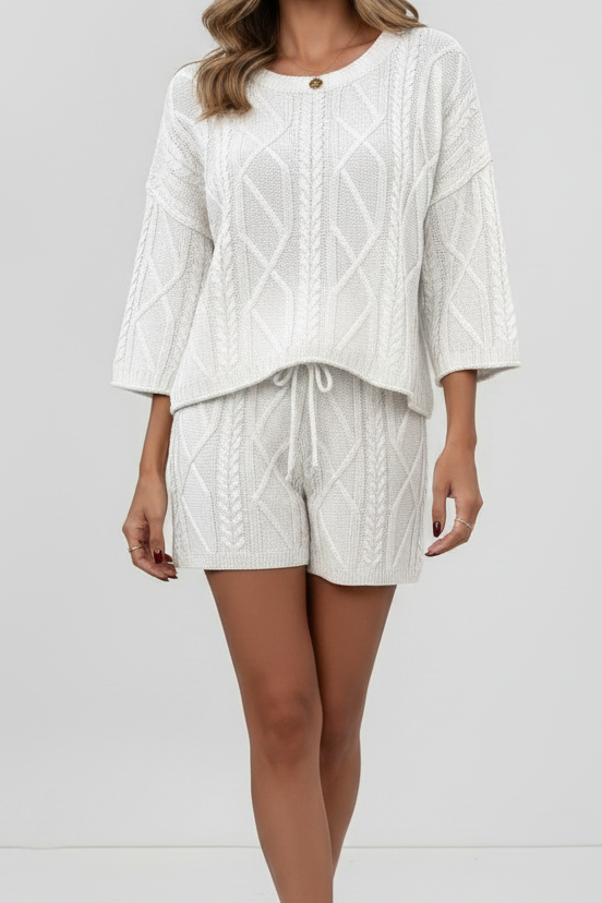 A woman modeling a matching two-piece knit set in off-white, featuring a cropped three-quarter sleeve top and coordinating shorts, shot against a seamless light gray studio background.